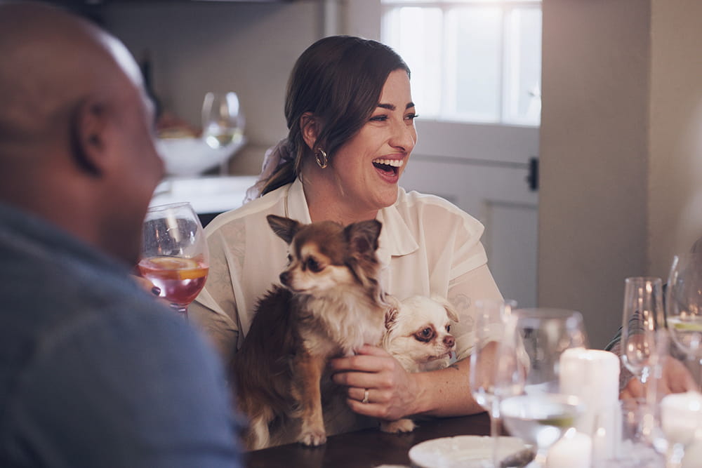 Woman holding two small dogs and glass of wine at dinner table